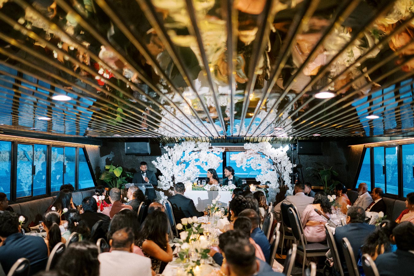 A luxurious wedding reception on a boat with guests seated around elegantly decorated tables. A speaker addresses the couple at a table adorned with white floral arrangements against a backdrop of large windows showcasing a scenic water view. The ceiling reflects the venue's festive ambiance.