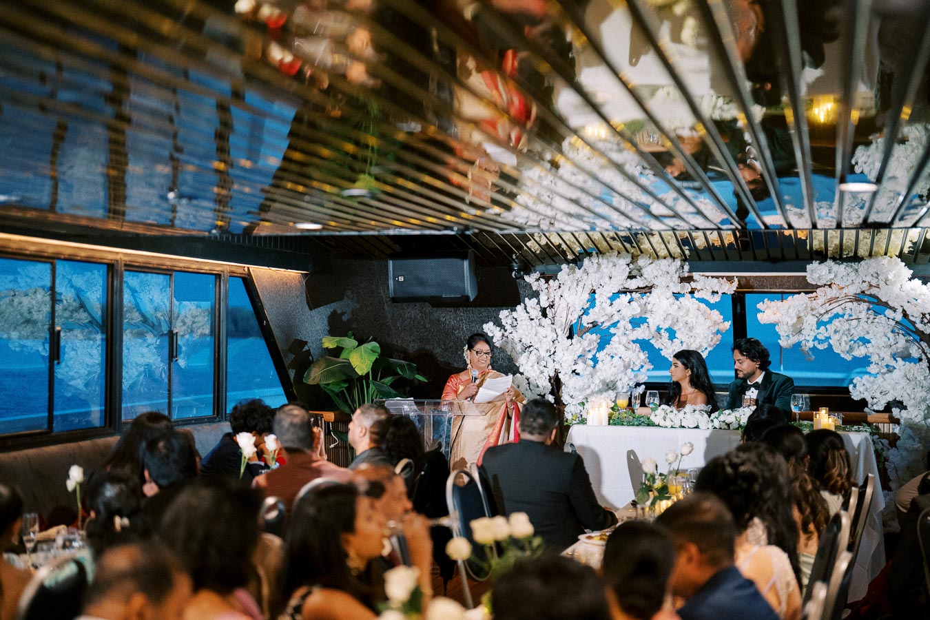 Elegant wedding reception on a boat with guests seated around tables, as a woman gives a speech. The couple sits at a decorated table with white floral arrangements and candles, overlooking a scenic water view through large windows.