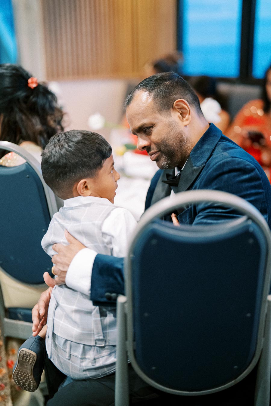 A father in a blue suit tenderly embracing his young son at a family event, surrounded by people seated on chairs.