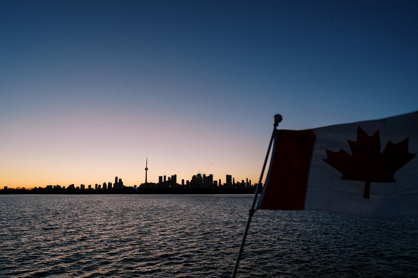 Sunset view of Toronto skyline across Lake Ontario with the Canadian flag in the foreground.