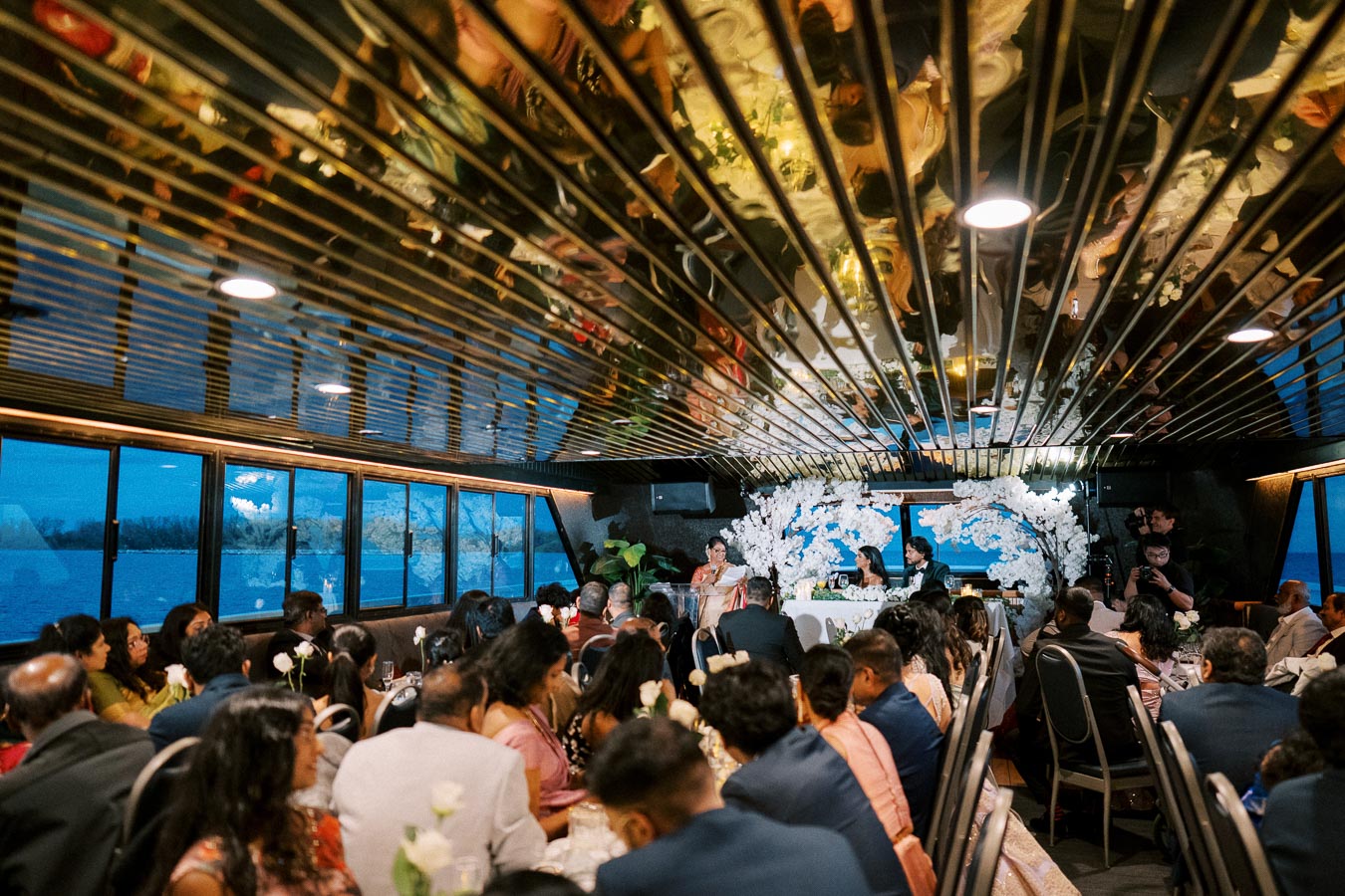 Wedding reception on a boat with guests seated at tables under a reflective ceiling. A beautifully decorated table with floral arrangements is at the front, set against large windows showcasing an evening water view.