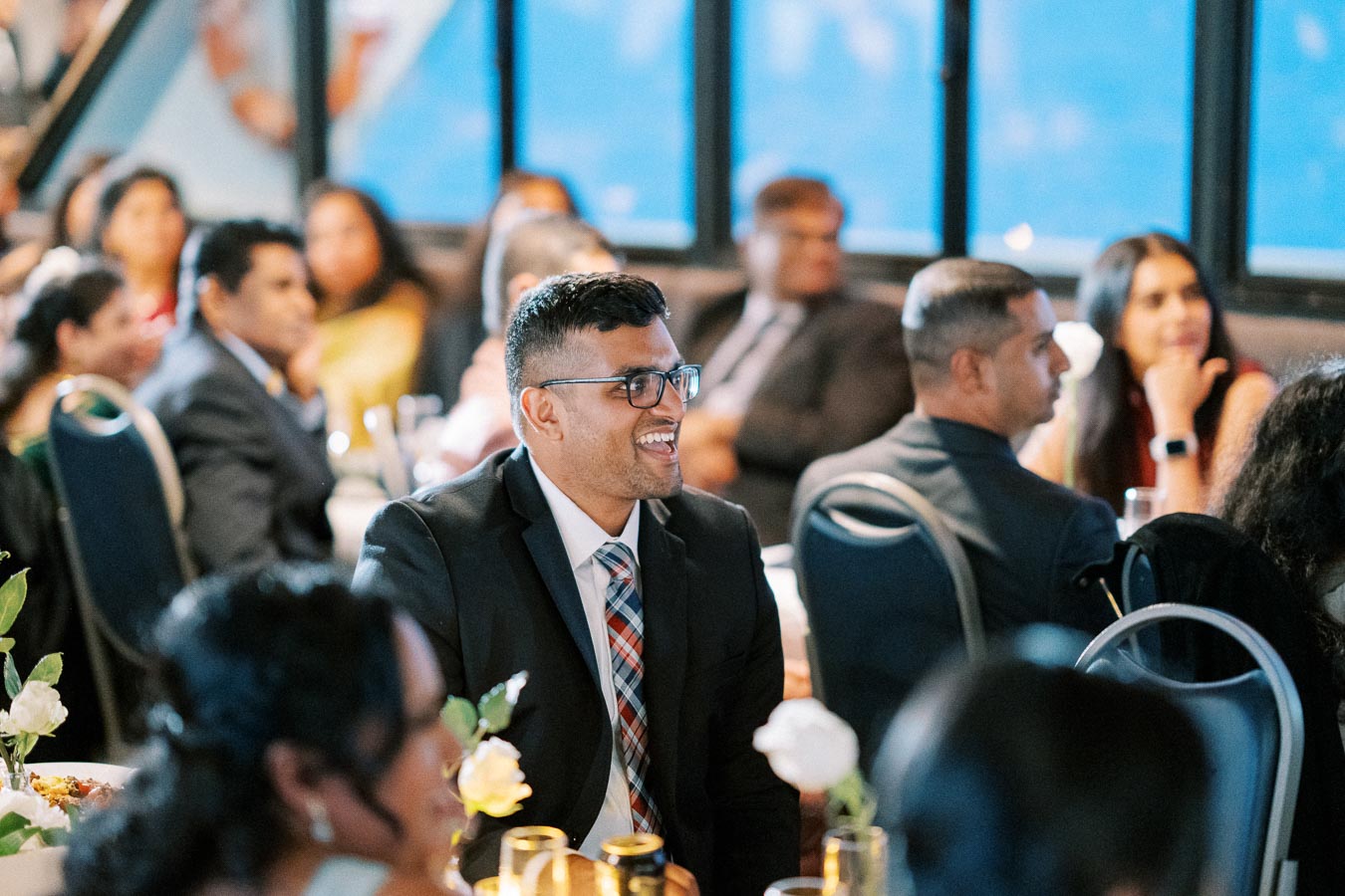 A man in a suit with glasses smiling at a formal event, surrounded by other well-dressed attendees seated at tables decorated with flowers.