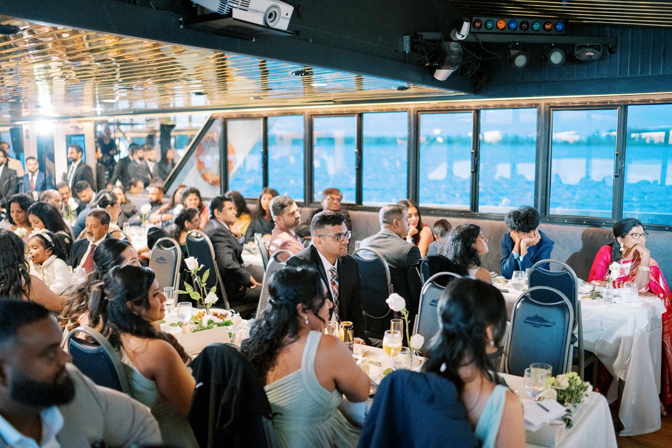 A diverse group of people attentively gathered at a formal event on a boat, seated around tables decorated with white flowers and glassware, with large windows providing a scenic view of the water.