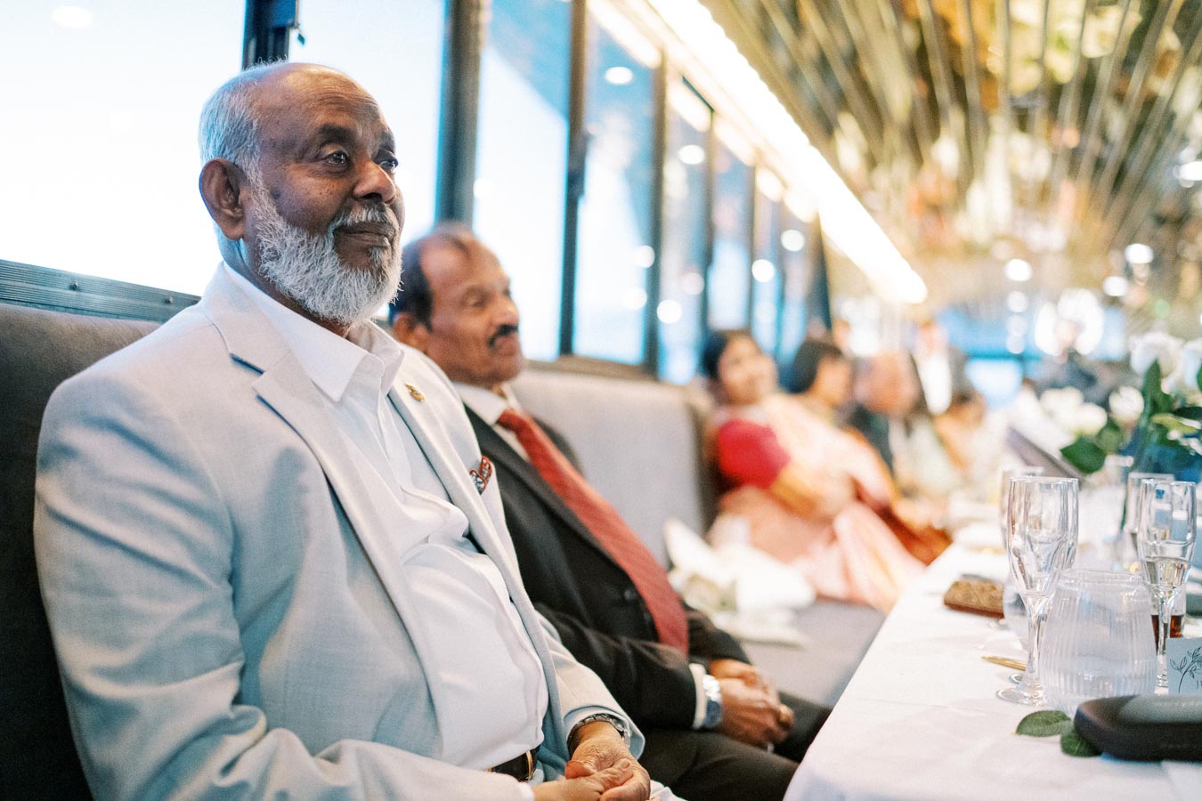 A group of elegantly dressed people sitting at a formal event inside a luxurious venue. The atmosphere is sophisticated with people engaged in conversation at a table adorned with glassware and floral decorations.