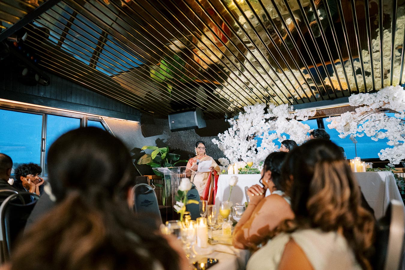 A person in a formal setting is delivering a speech at an elegant event, with a decorative floral backdrop and attentive audience members in the foreground, creating a celebratory atmosphere.