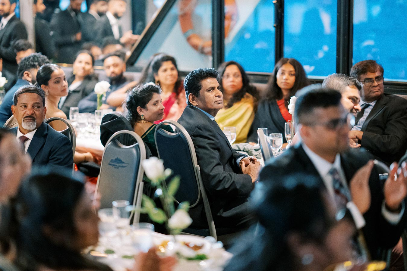 Formal event with guests seated at tables, attentively listening, dressed in suits and traditional attire, in a warmly lit banquet hall setting.