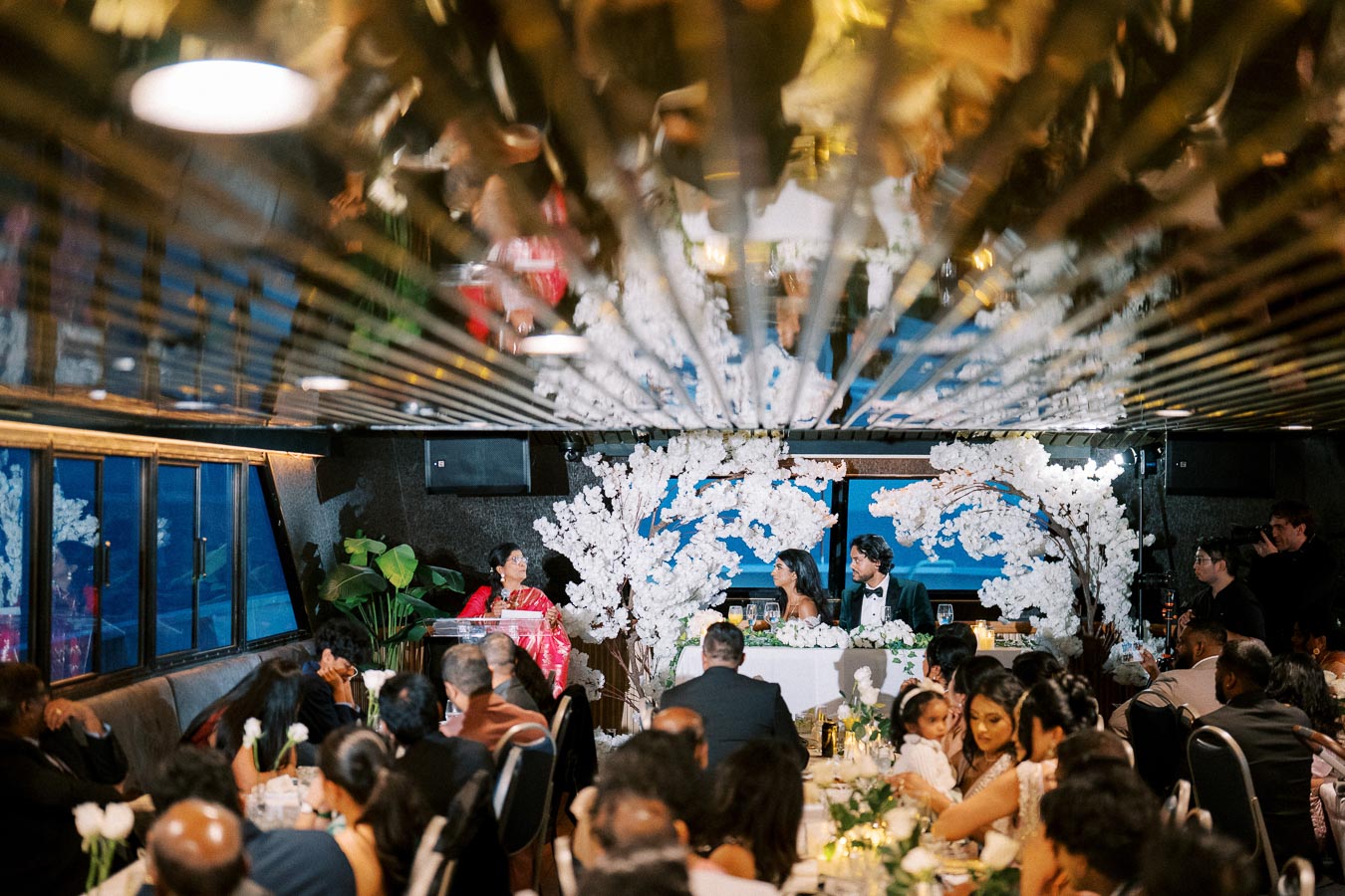 Wedding reception on a boat with elegant floral decorations, a beautifully set table, and guests listening to a speech under a reflective ceiling.