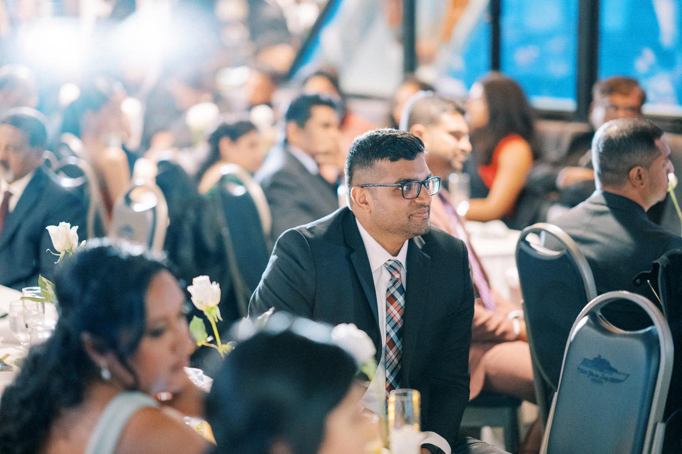 A man in a suit and glasses sitting at a formal event, surrounded by other attendees wearing evening attire, with white rose centerpieces on the tables and soft lighting in the background.