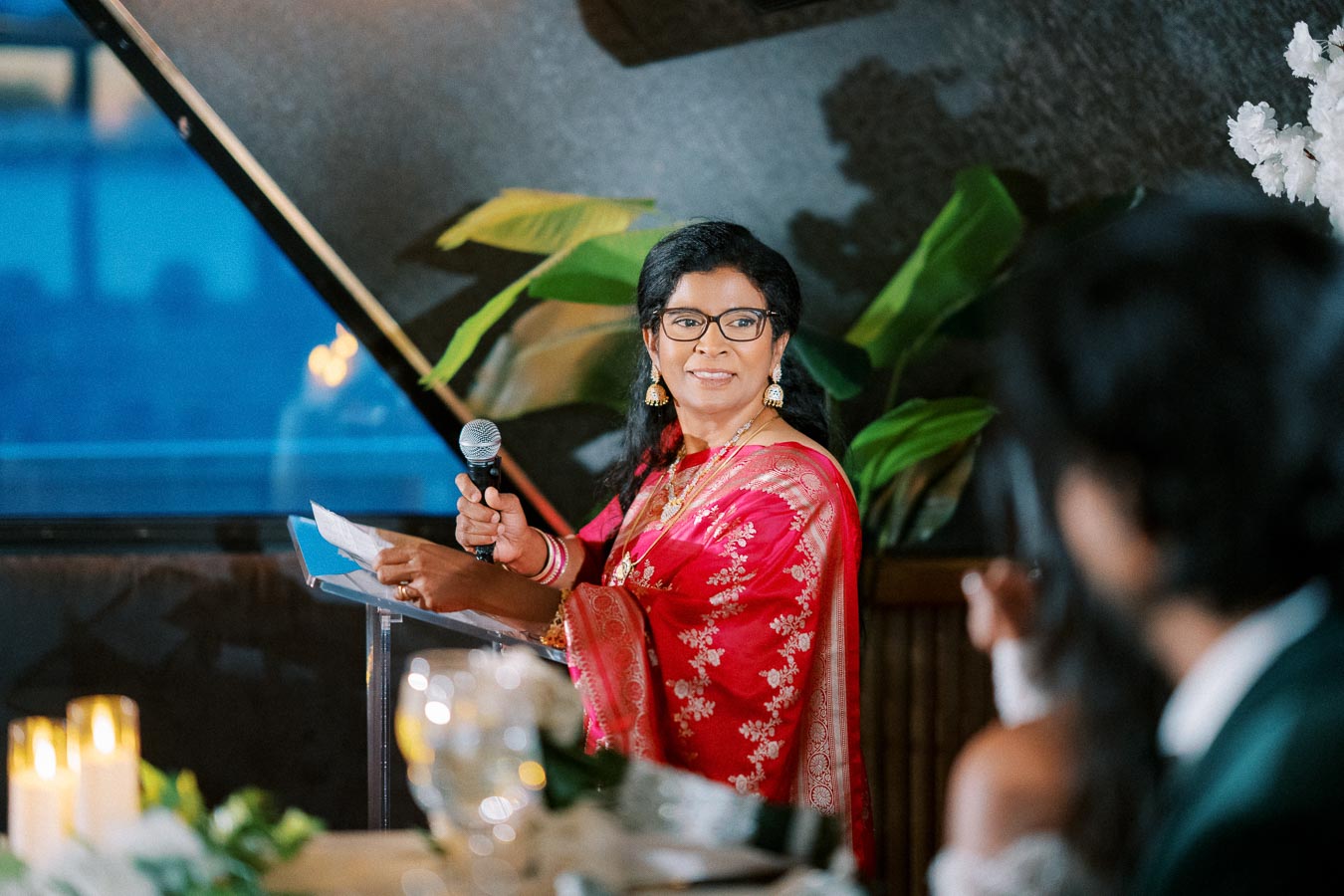 A woman in a red floral saree speaks into a microphone at an indoor event. She stands at a podium, surrounded by candles and green plants, with a blurred audience member in the foreground.
