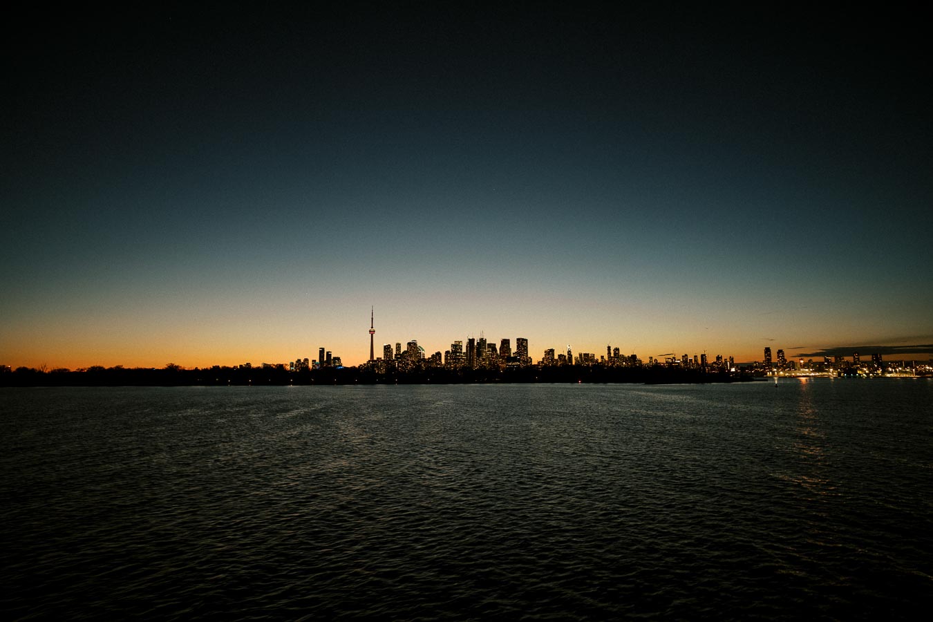Toronto skyline at dusk with clear sky and illuminated buildings reflected on Lake Ontario.