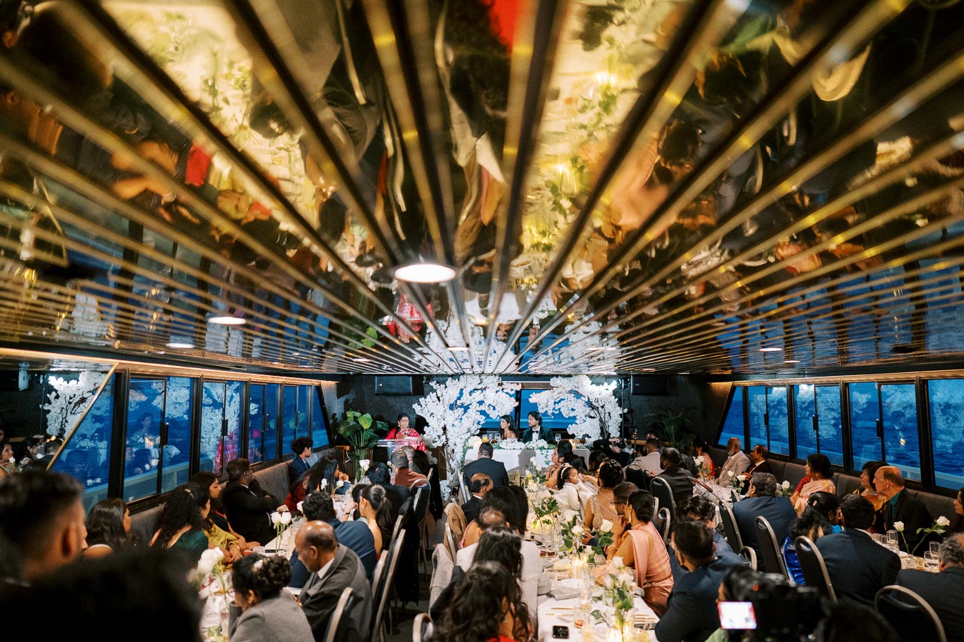 Elegant wedding reception on a boat with guests seated at decorated tables, blue ocean visible through large windows, and floral arrangements adorning the venue.