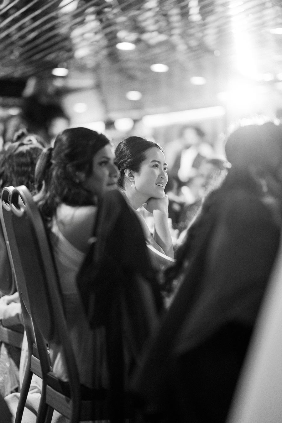 A black and white photograph of a woman smiling while sitting among a group of people during an indoor event, with a blurred, illuminated background.