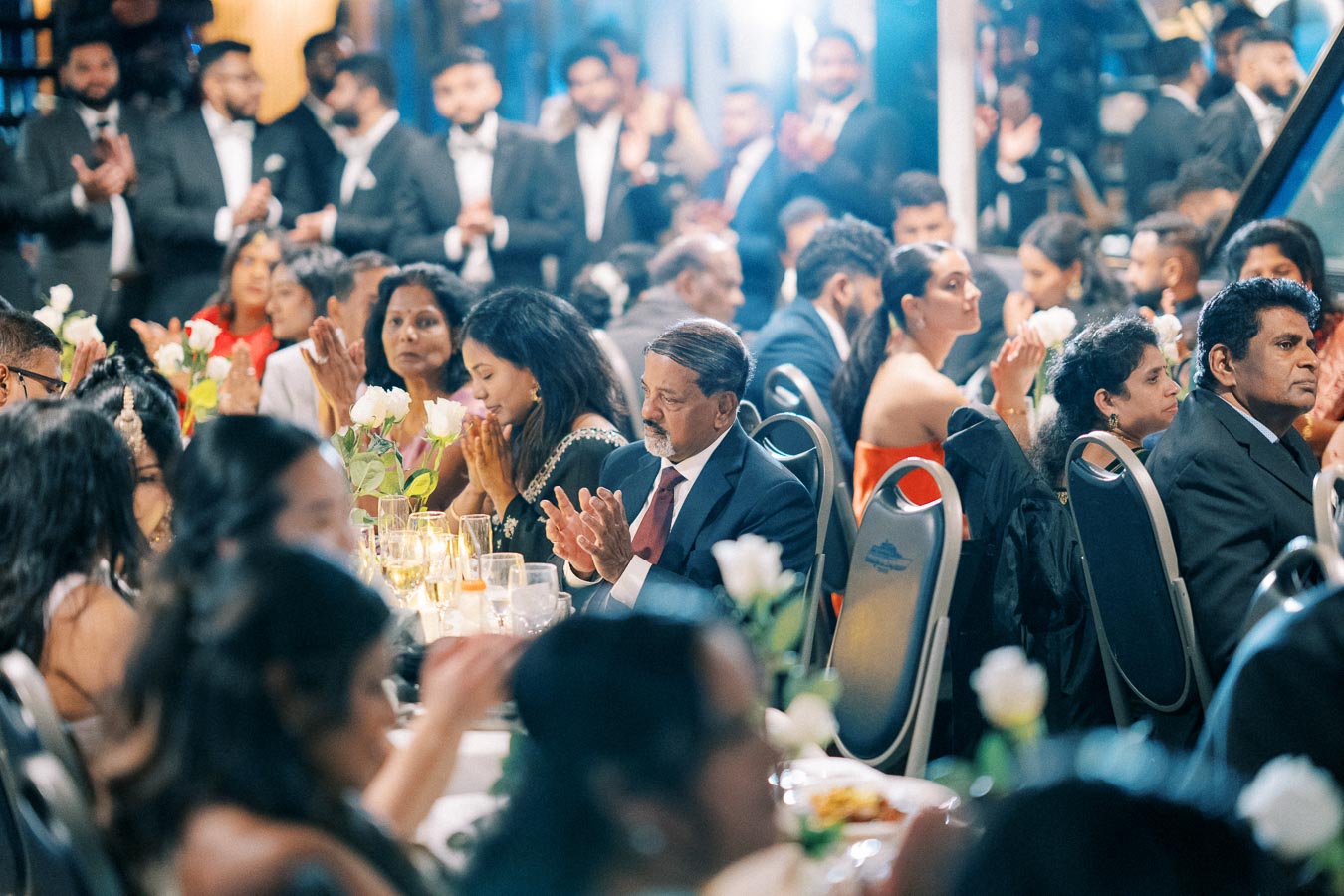 Elegant banquet hall with guests sitting at a formally set table decorated with white roses, clapping and enjoying an event. Attendees are dressed in formal attire, creating a sophisticated atmosphere.