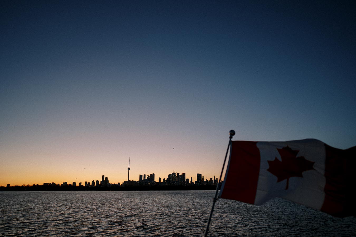 Sunrise over Toronto skyline with Canadian flag in foreground across calm lake waters.