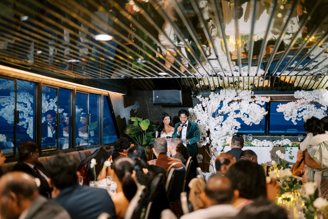 A couple giving a heartfelt speech at their wedding reception, surrounded by elegant white floral decorations and guests seated at tables.