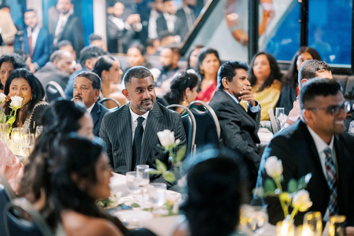 A well-dressed group of people attentively listening at an elegant event or conference, seated around tables decorated with flowers and candles, creating a sophisticated atmosphere.