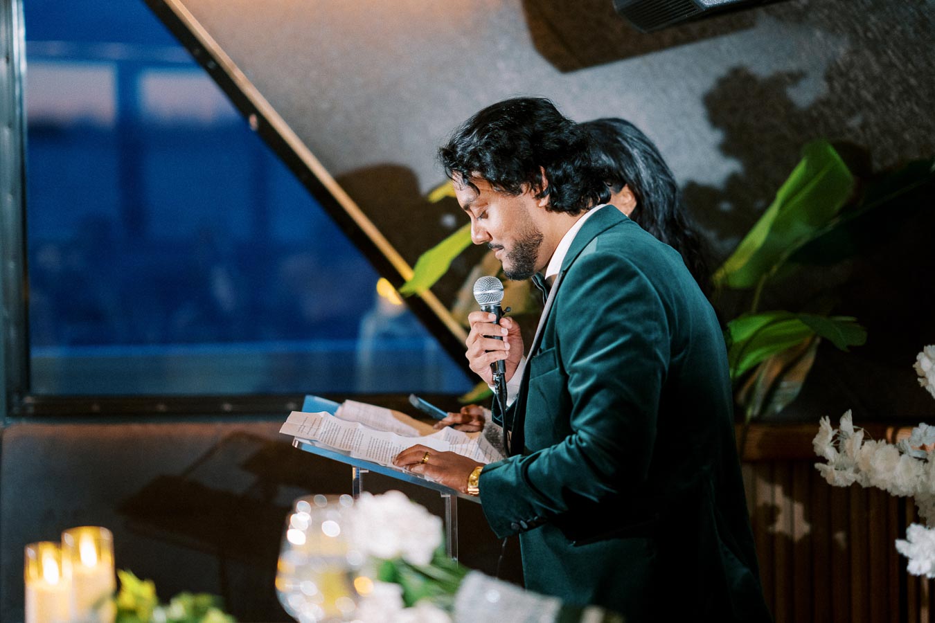 Man in a dark suit giving a speech at an indoor event, holding a microphone and reading from a paper, with blurred candles and flowers in the foreground.