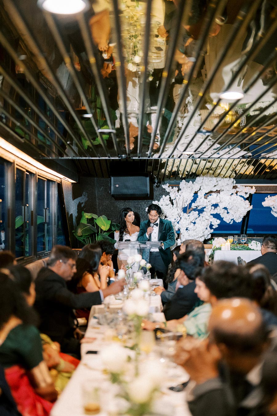 A couple giving a speech at a wedding reception, with guests seated at a long, elegantly decorated table featuring white roses. The room has a modern design with a reflective ceiling and large windows offering a nighttime view.