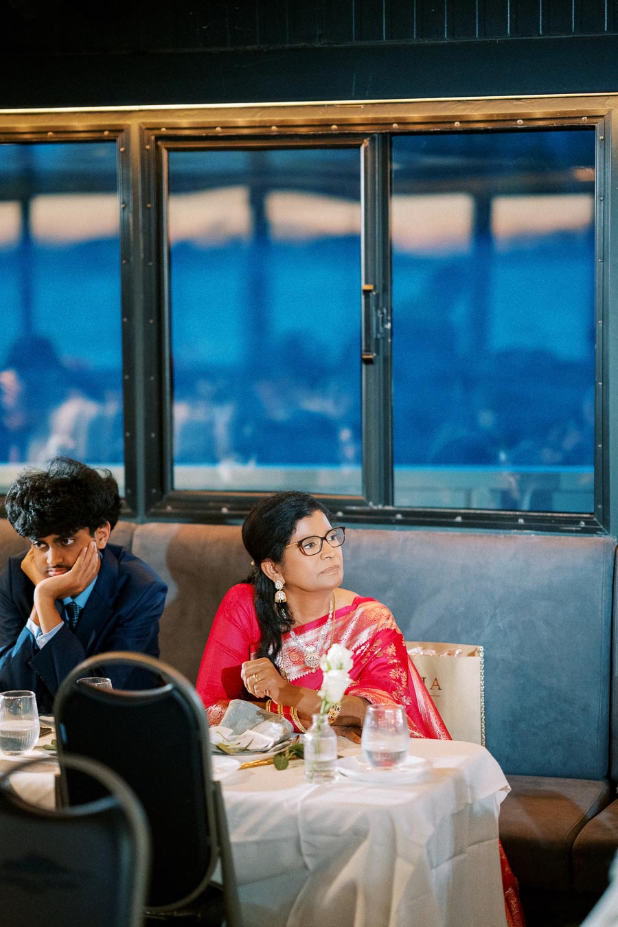 A man in a suit and a woman in a traditional red sari seated at a dining table, attentively listening during an evening event in a room with large windows.