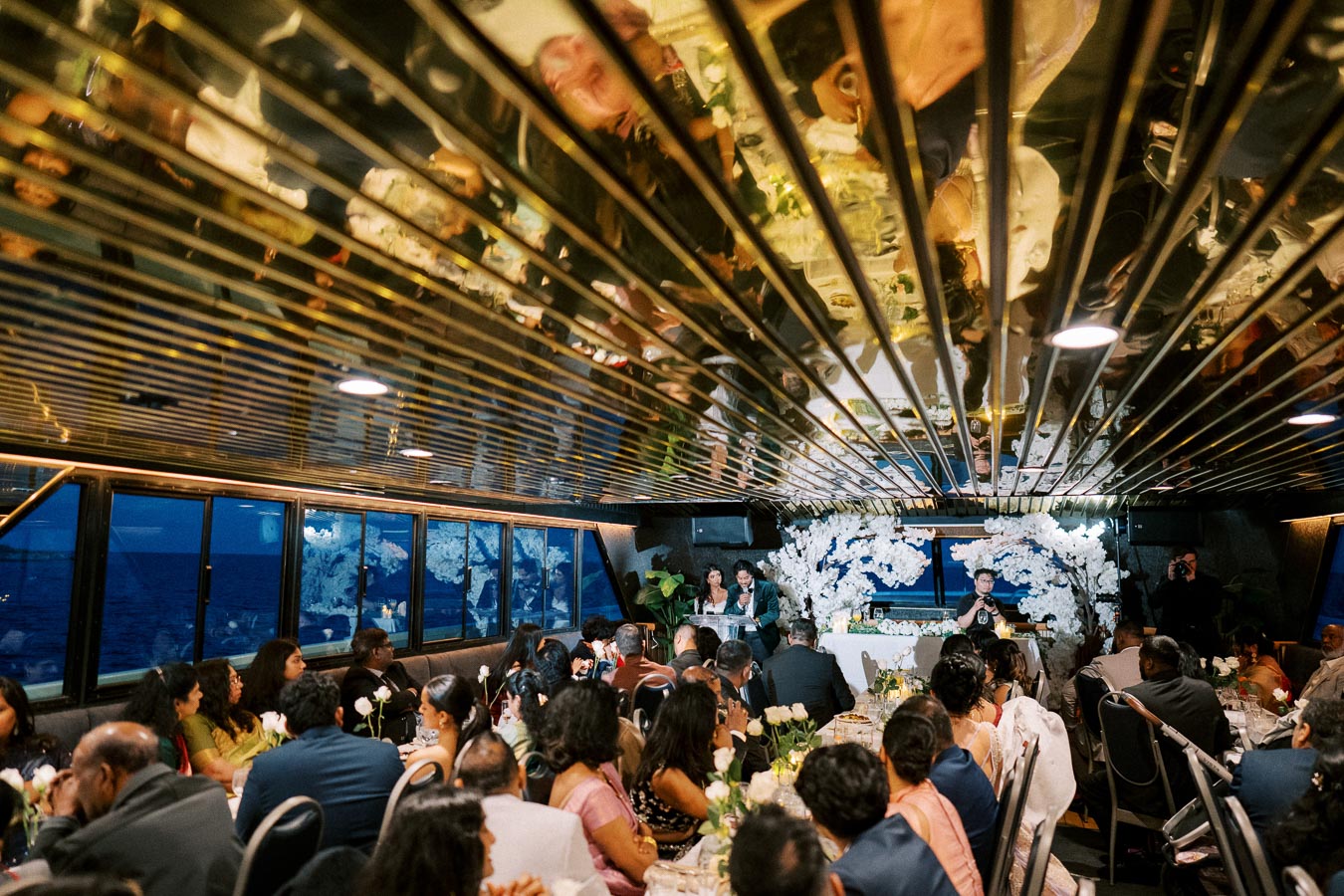 Elegant wedding reception on a boat with guests seated at tables, beautifully decorated with white floral arrangements and a reflective ceiling, set against an ocean backdrop.