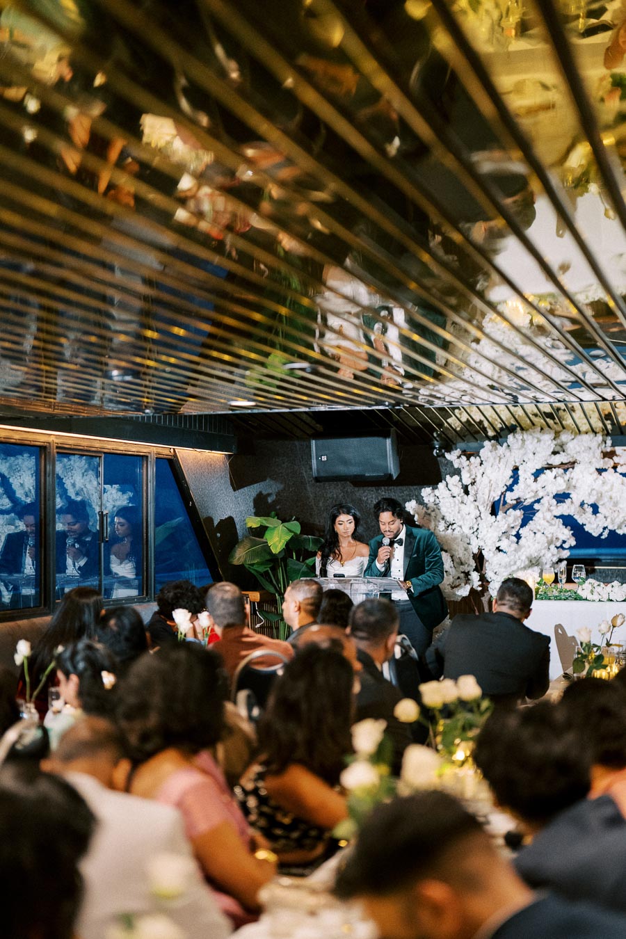 A bride and groom giving a speech in a beautifully decorated wedding venue with guests seated at elegantly arranged tables, adorned with white flowers and ambient lighting.