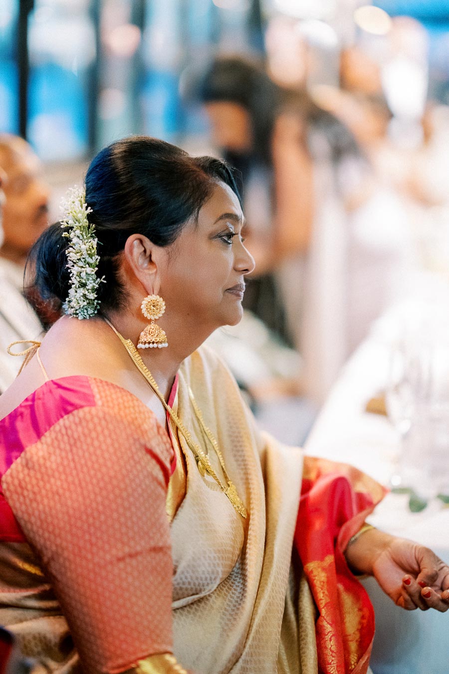 A woman in a traditional silk sari with red and gold accents is sitting indoors, wearing intricate gold earrings and a necklace. Her hair is elegantly adorned with small white flowers.