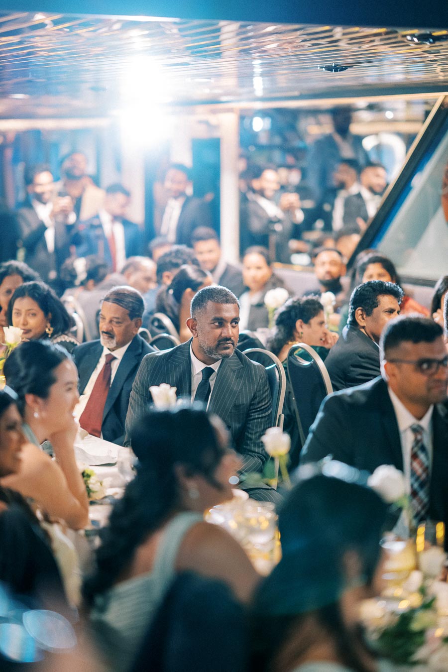 A formal gathering with elegantly dressed guests seated at tables, engaged in attentive listening, adorned with flowers and soft lighting in a sophisticated indoor setting.