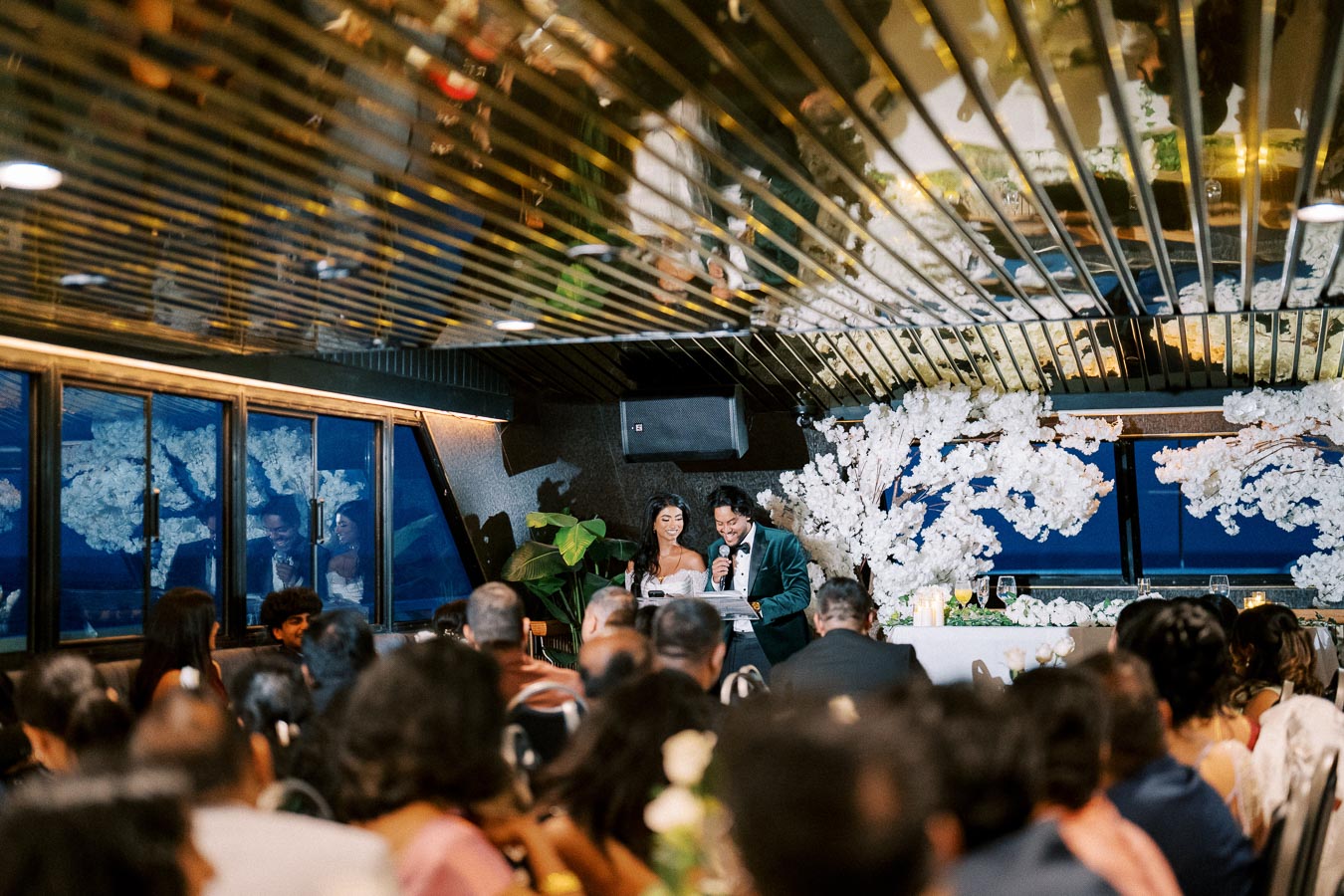 A couple giving a wedding speech on a decorated stage with white floral arrangements in an elegant venue, surrounded by guests.