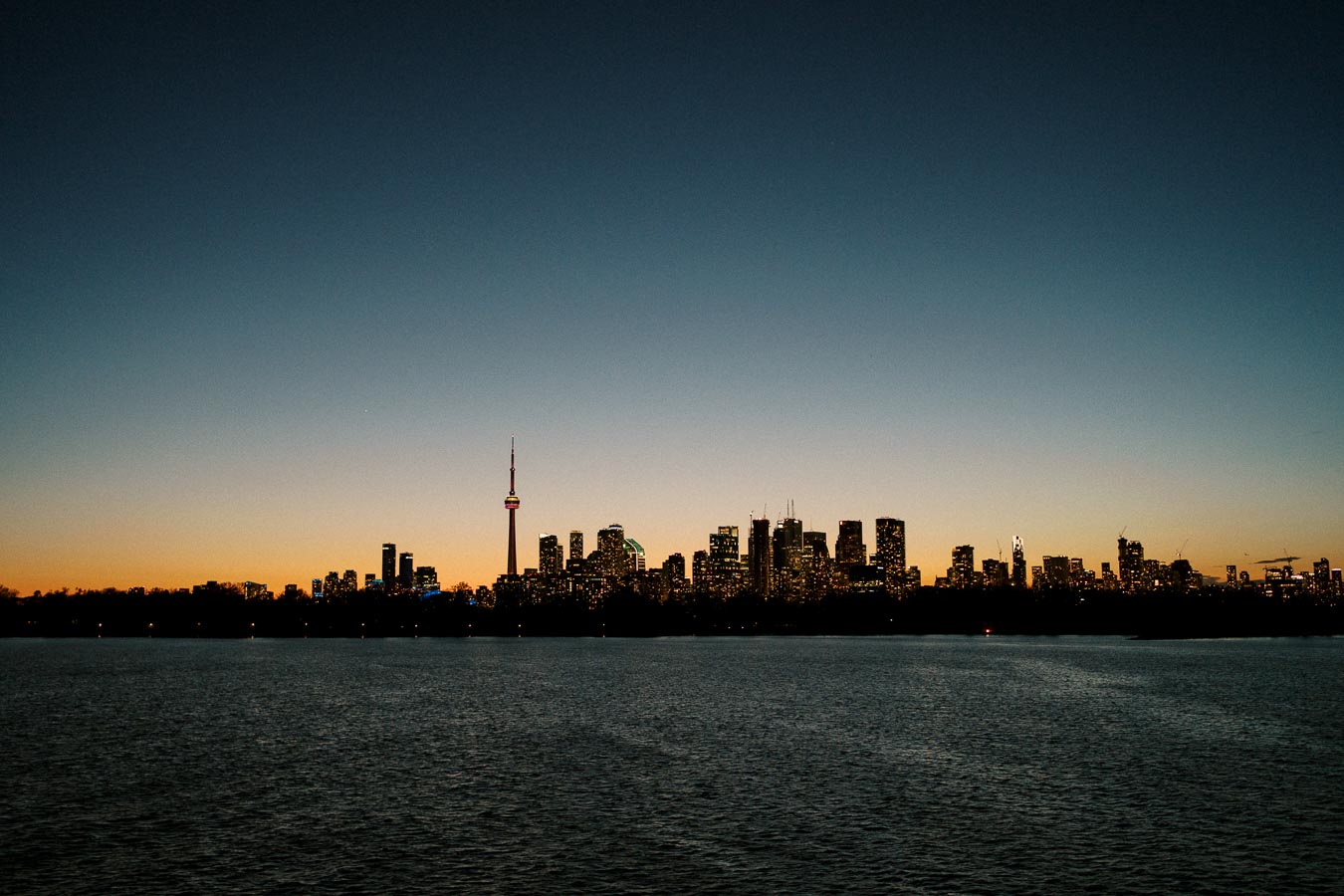 Skyline of Toronto at dusk with the CN Tower prominently featured, city lights reflecting over Lake Ontario under a clear evening sky.