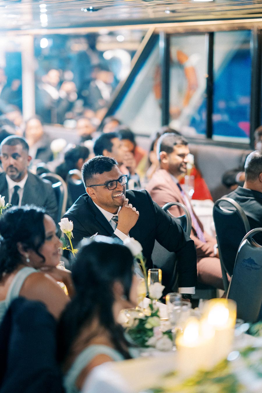 Group of people enjoying a formal event, seated at a decorated table with flowers and candles, with one man smiling and engaging with the speaker