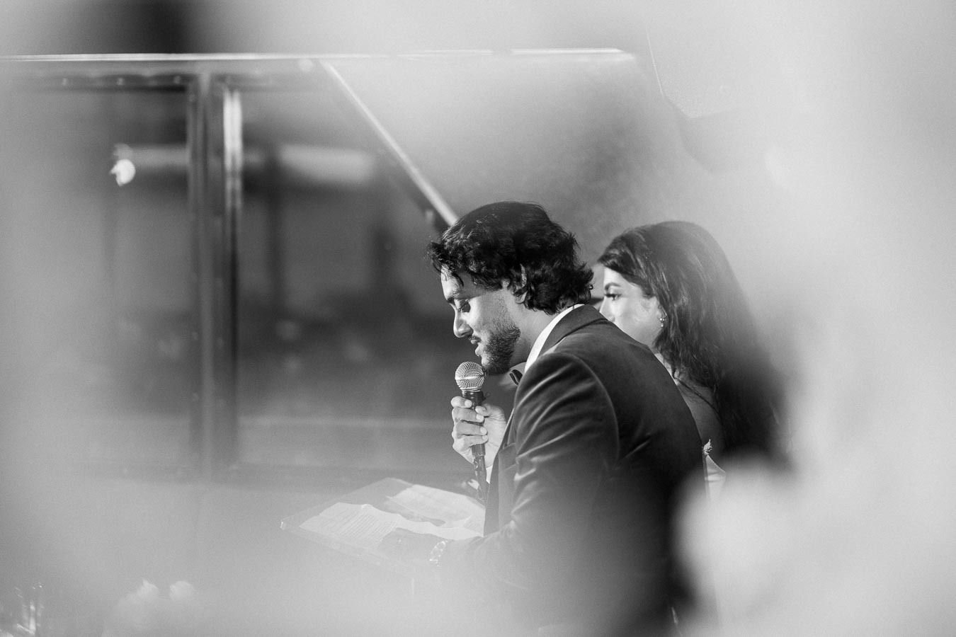 Black and white image of a man speaking into a microphone, with a woman standing beside him, both in formal attire, in a softly blurred setting.