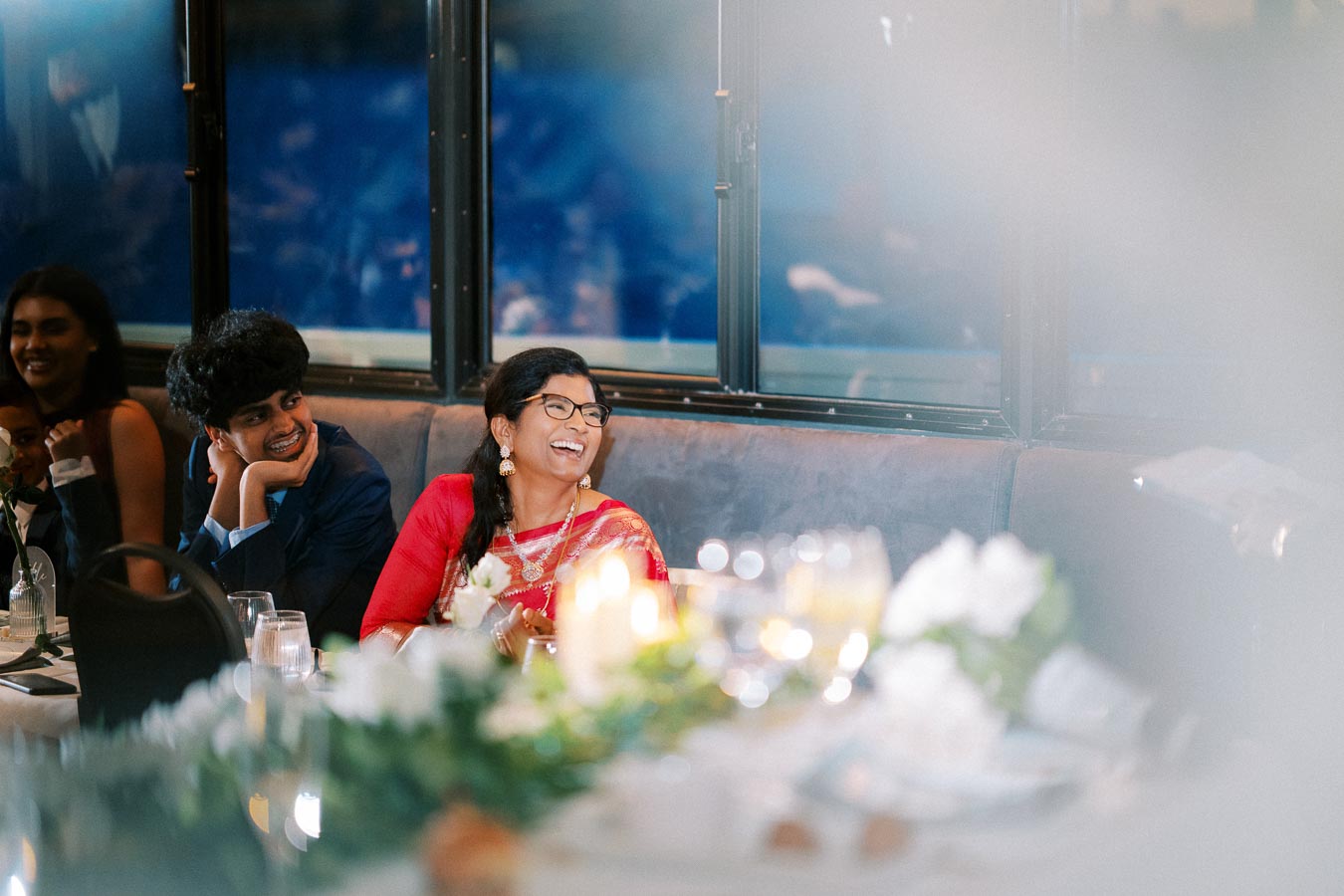 A group of people laughing and enjoying a festive gathering, seated at a decorated table with soft lighting and elegant floral arrangements.