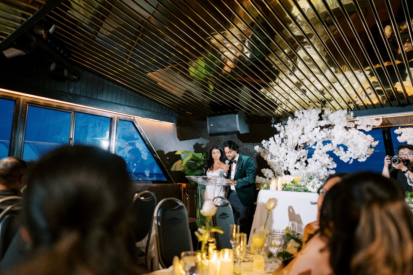 Couple giving a wedding speech in an elegant venue with floral decorations and guests seated at tables.