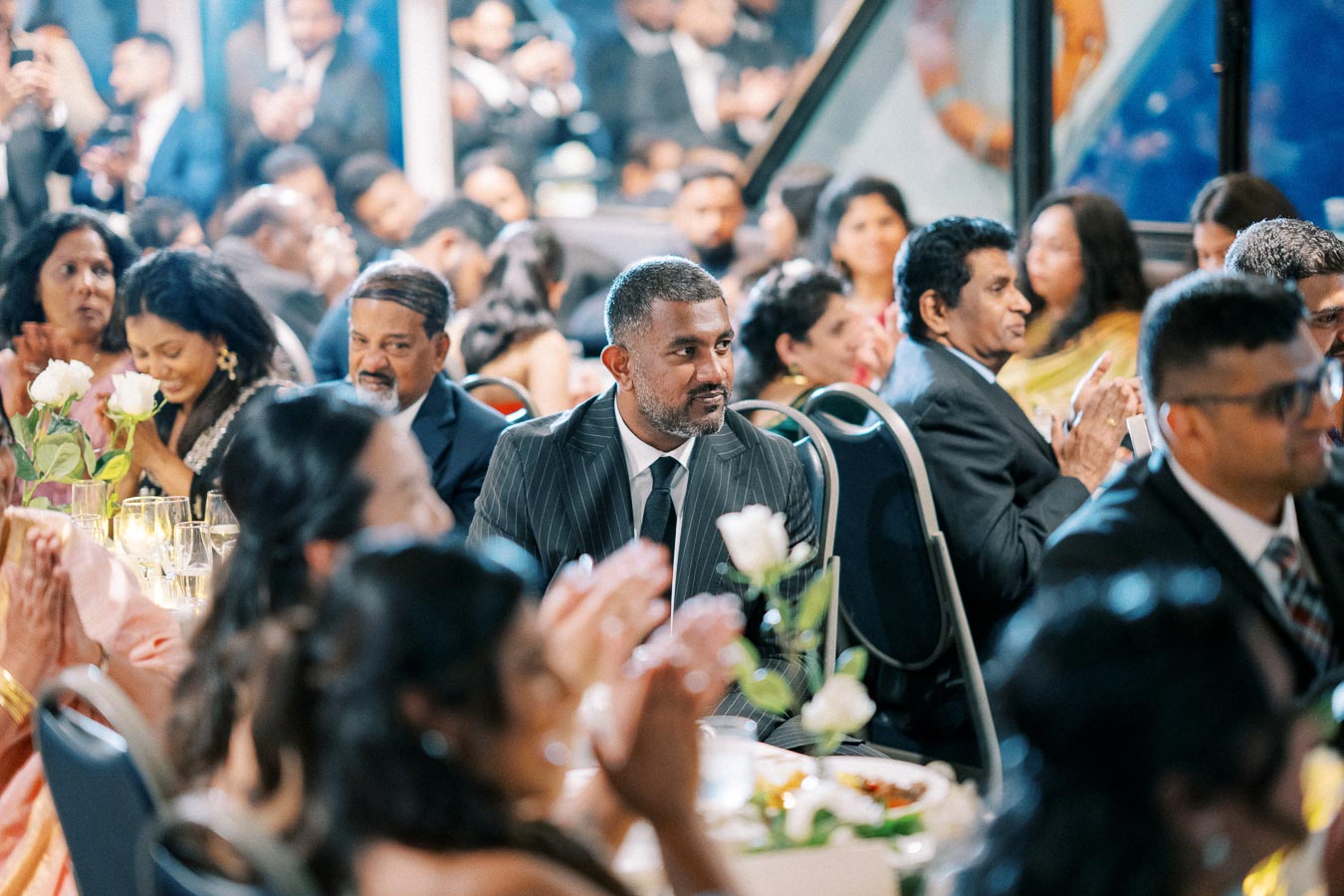A diverse group of well-dressed individuals sitting at a formal event, attentively listening and clapping, with elegant table settings and floral arrangements.
