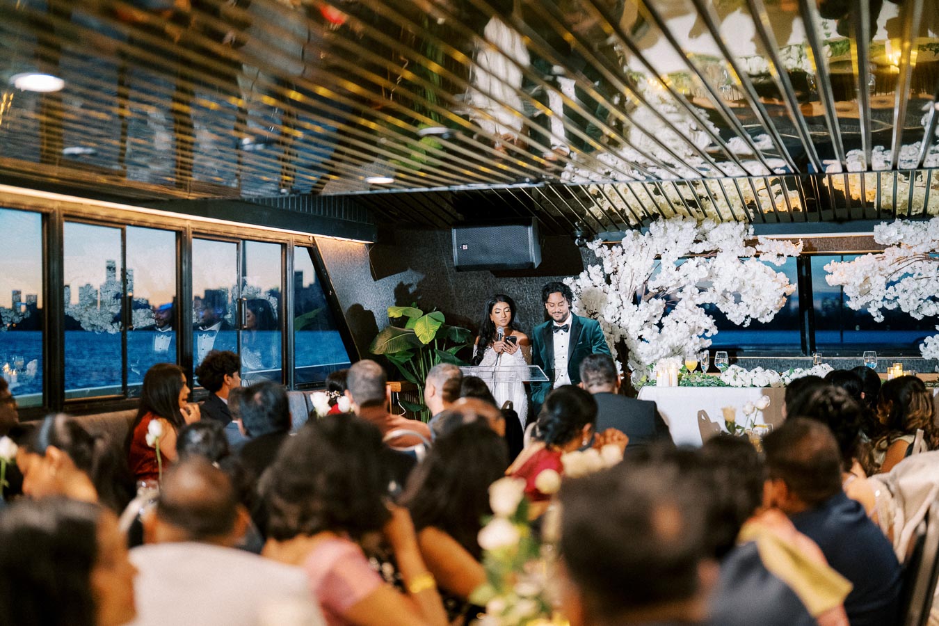 A couple giving a speech at a wedding reception on a boat, surrounded by guests. The venue features elegant floral decorations and large windows with a view of the city skyline and waterfront.