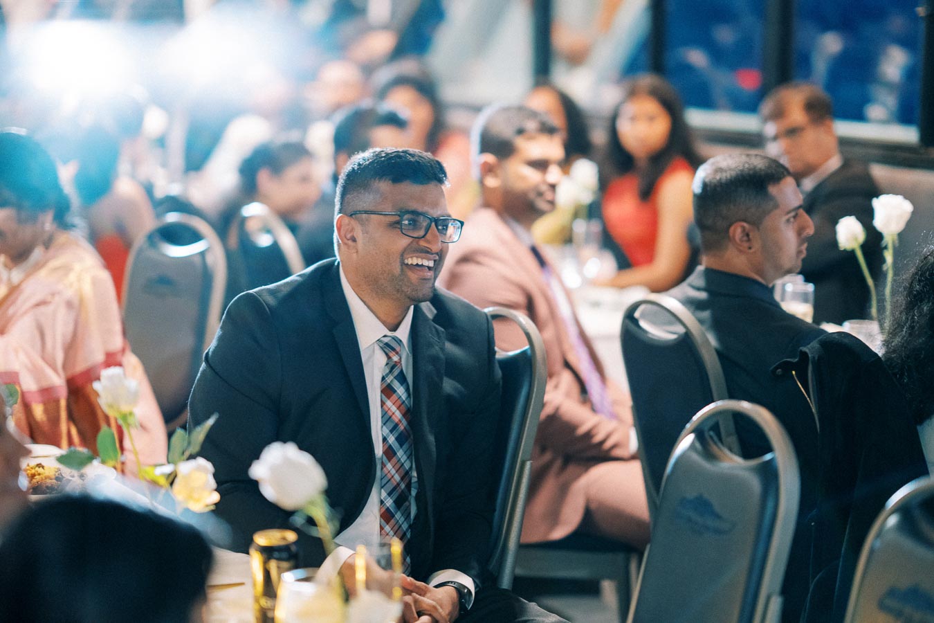 A group of elegantly dressed people sitting at a formal event, with a smiling man in a suit and tie in focus, surrounded by tables and white flower centerpieces.