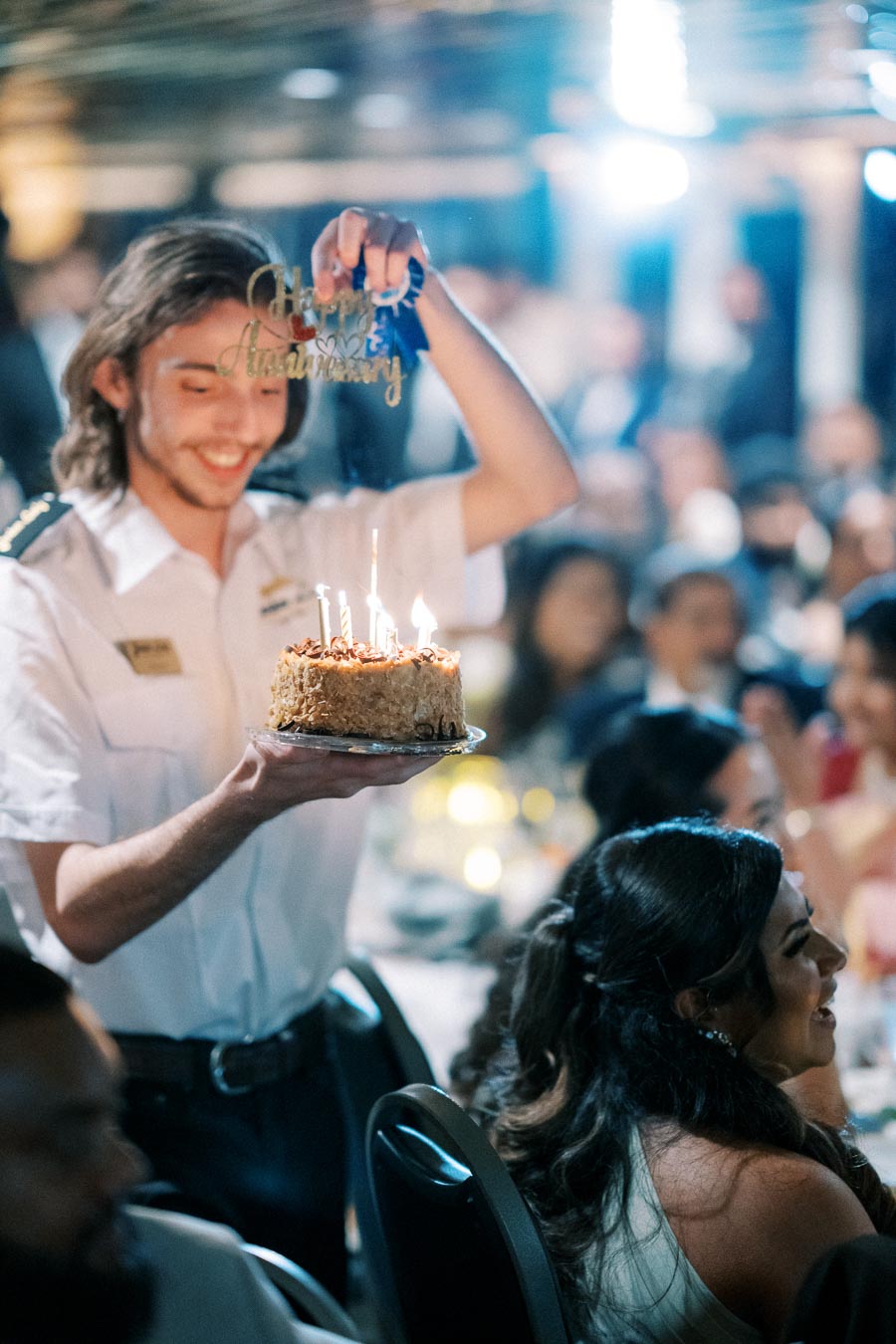 Waiter holding a lit anniversary cake at a celebratory event with smiling guests