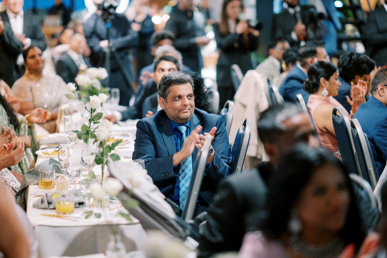 A well-dressed man in a suit clapping at a formal event, surrounded by elegantly dressed guests seated at tables adorned with white roses and glassware.