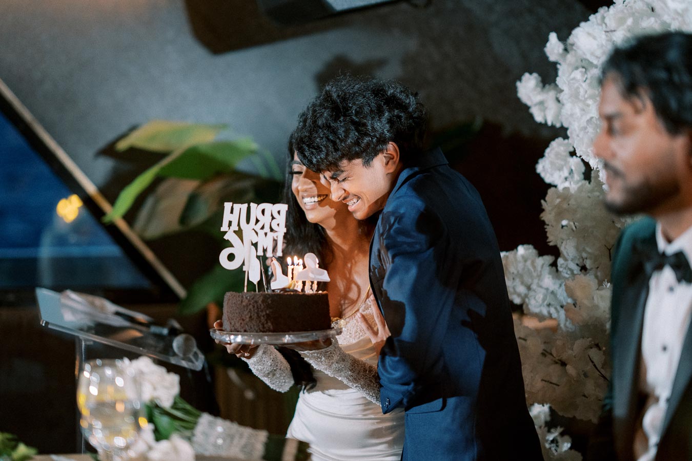 A couple happily embraces while holding a lit birthday cake adorned with creative decorations, surrounded by elegant party decor in a festive setting.