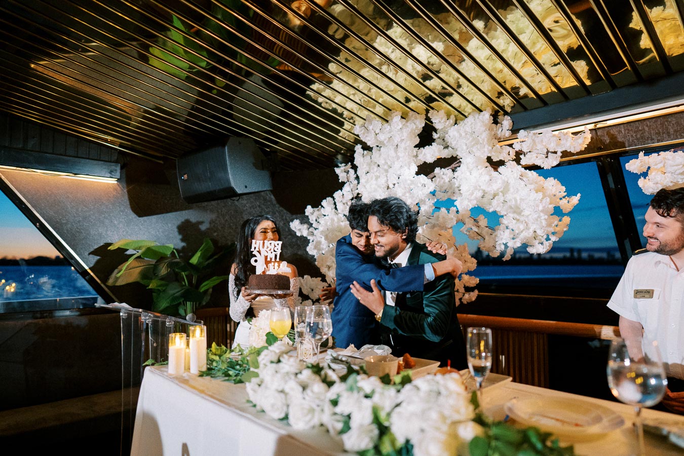Celebration aboard a scenic sailboat with individuals embracing, surrounded by elegant floral decorations and decor, a woman holding a cake with a lettered topper, and a city skyline visible in the background at dusk.