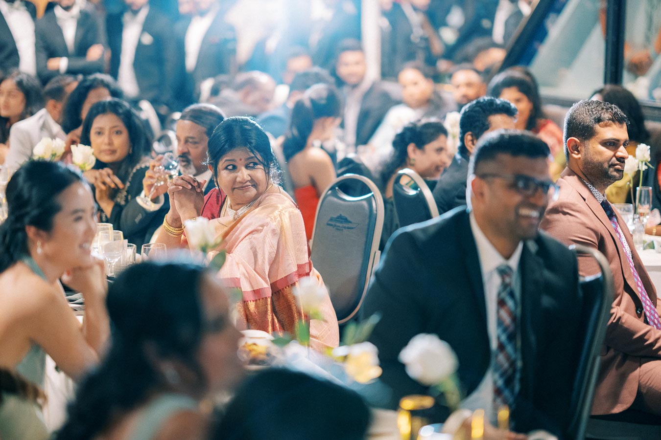 A lively wedding reception with guests seated at tables, including a woman in a pink saree enjoying the event, surrounded by elegantly dressed attendees.