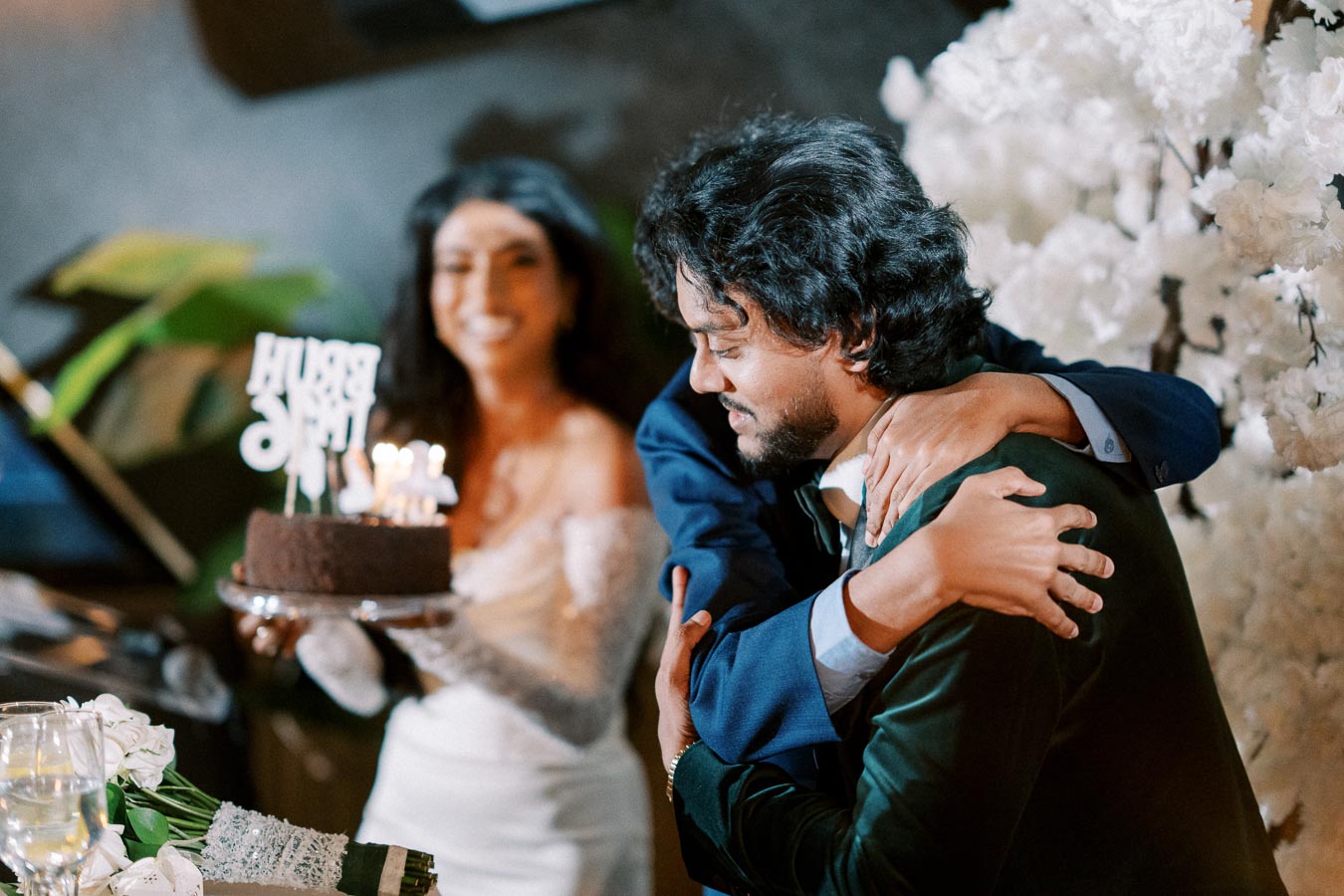 Two men share a heartfelt hug at a celebration, with a woman in the background holding a lit birthday cake. The room is elegantly decorated with flowers and champagne glasses, creating a festive atmosphere.