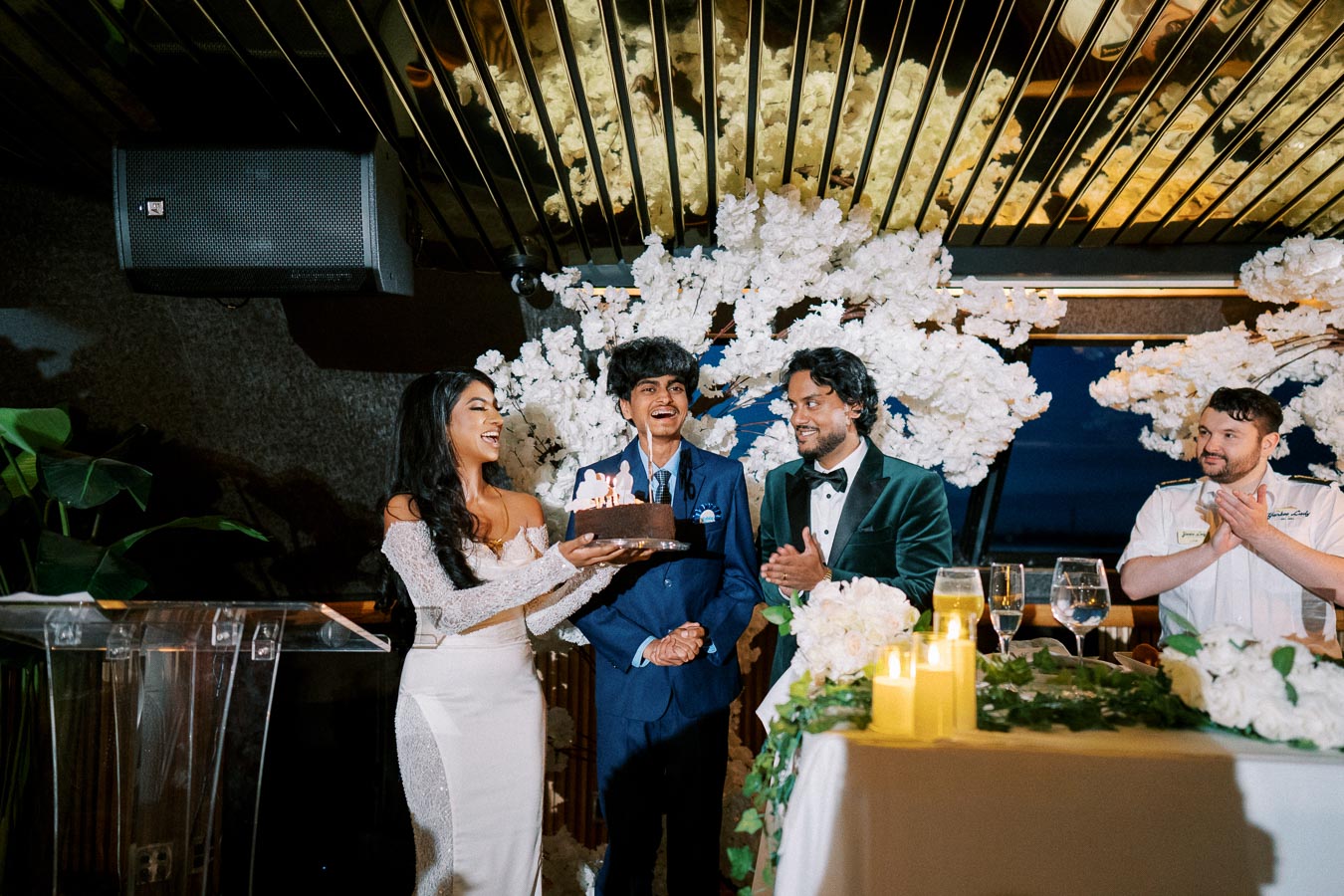 A woman in a white dress presenting a lit birthday cake to a young man in a blue suit, surrounded by smiling people and floral decorations during an elegant celebration.