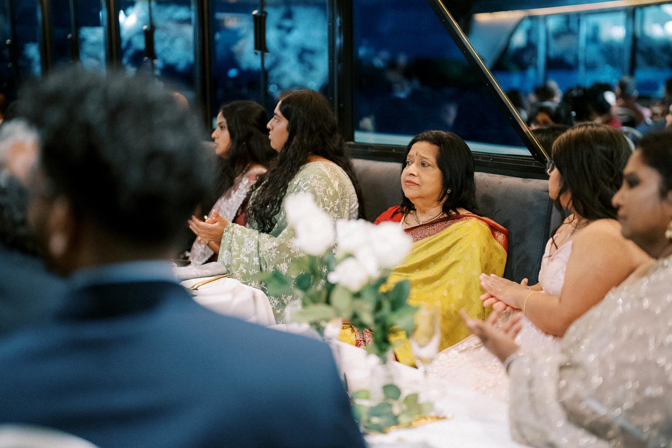 A group of people seated at a formal event, dressed in elegant attire, with a woman in a yellow saree as the focal point. The table is set with flowers and glassware, indicating a celebratory occasion in a sophisticated venue.
