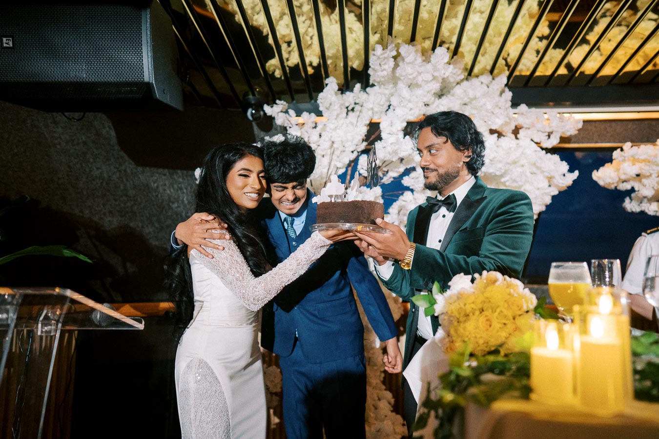A joyful celebration scene with a woman in a white dress embracing two men, one holding a chocolate cake, under a decorative floral arrangement.