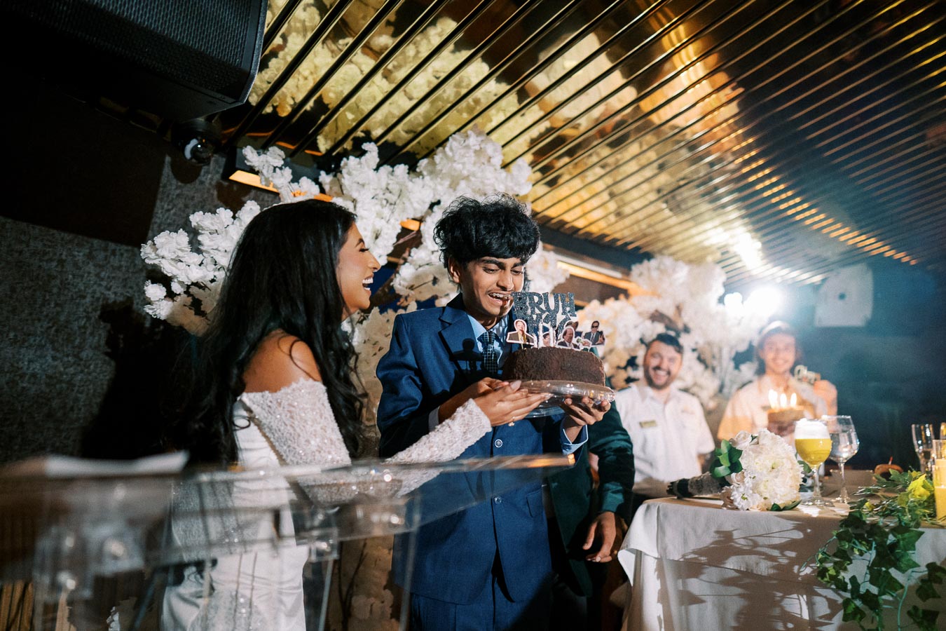 A joyous celebration scene with a young man in a blue suit holding a decorated chocolate cake, surrounded by smiling guests and white floral decorations, in a warmly lit venue.