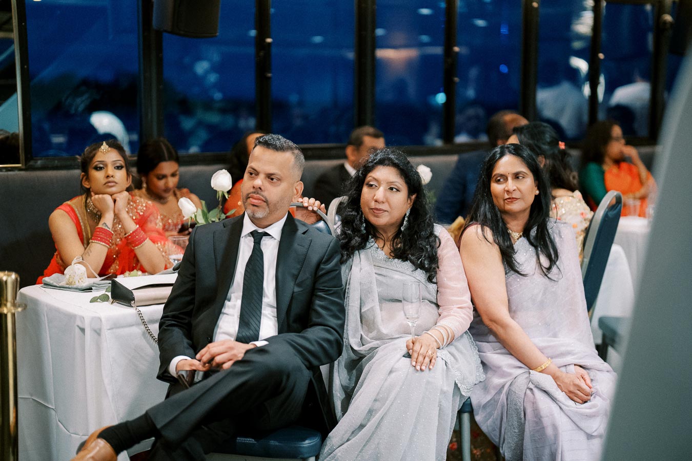 Group of people seated at a formal event, with a man in a suit and two women in elegant attire, paying attention to an unseen speaker.
