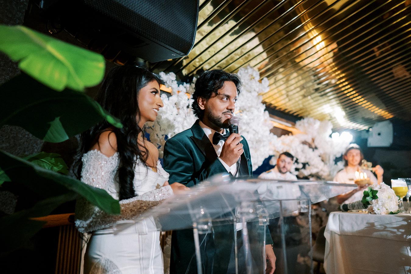 A couple stands at a lectern giving a speech during a wedding reception, with elegant floral decorations in the background. The bride is in an off-shoulder white dress, and the groom wears a dark suit with a bow tie.