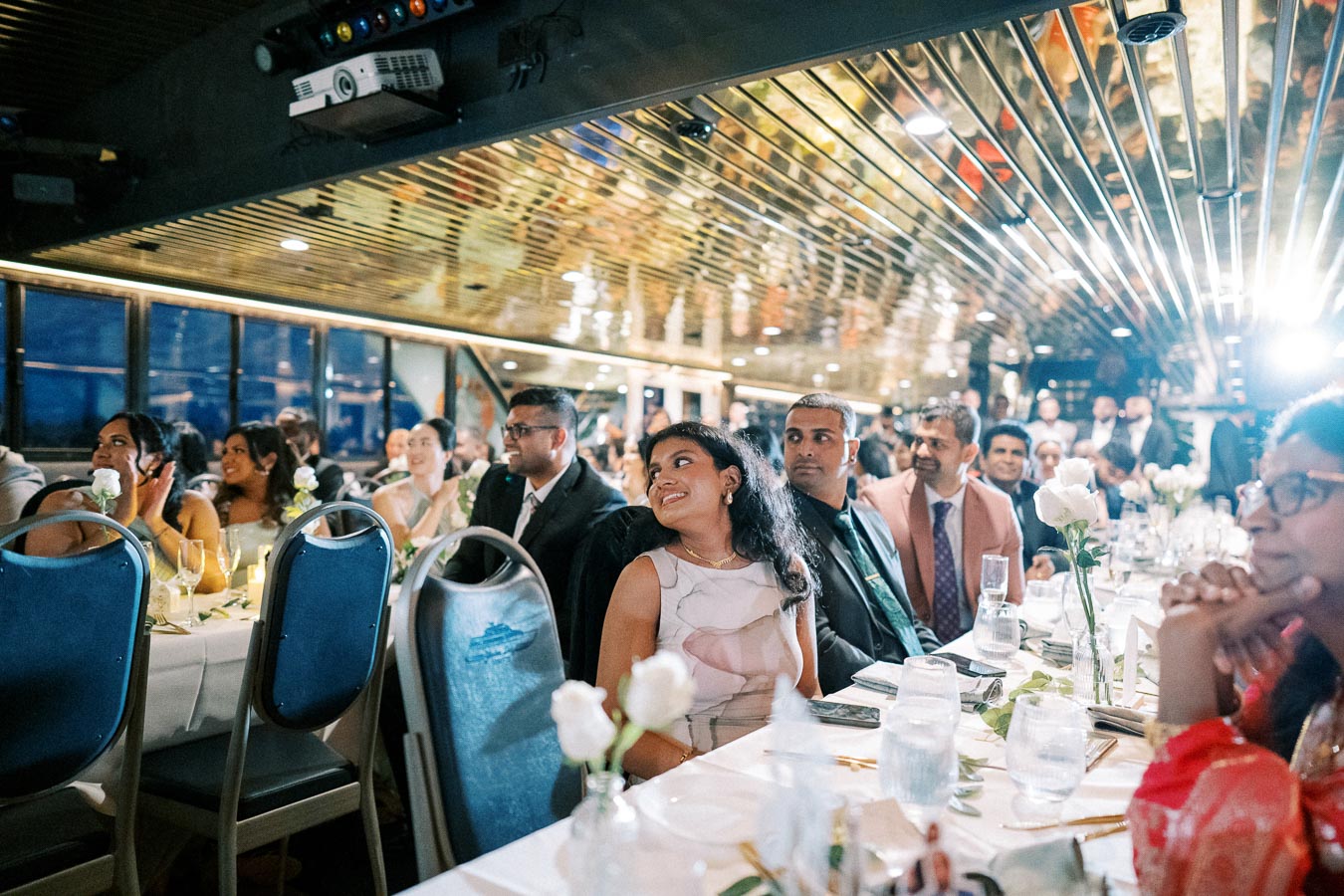 A lively group of elegantly dressed guests enjoying a formal dinner event aboard a luxury yacht, featuring decorative lighting and a beautifully set table with white roses and glassware.