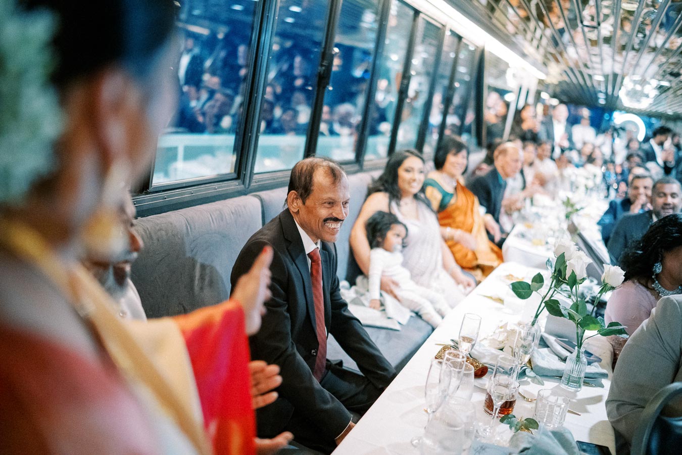 A group of people in formal attire sitting around a banquet table, smiling and engaged in conversation, with a festive atmosphere and floral decorations in a brightly lit room.